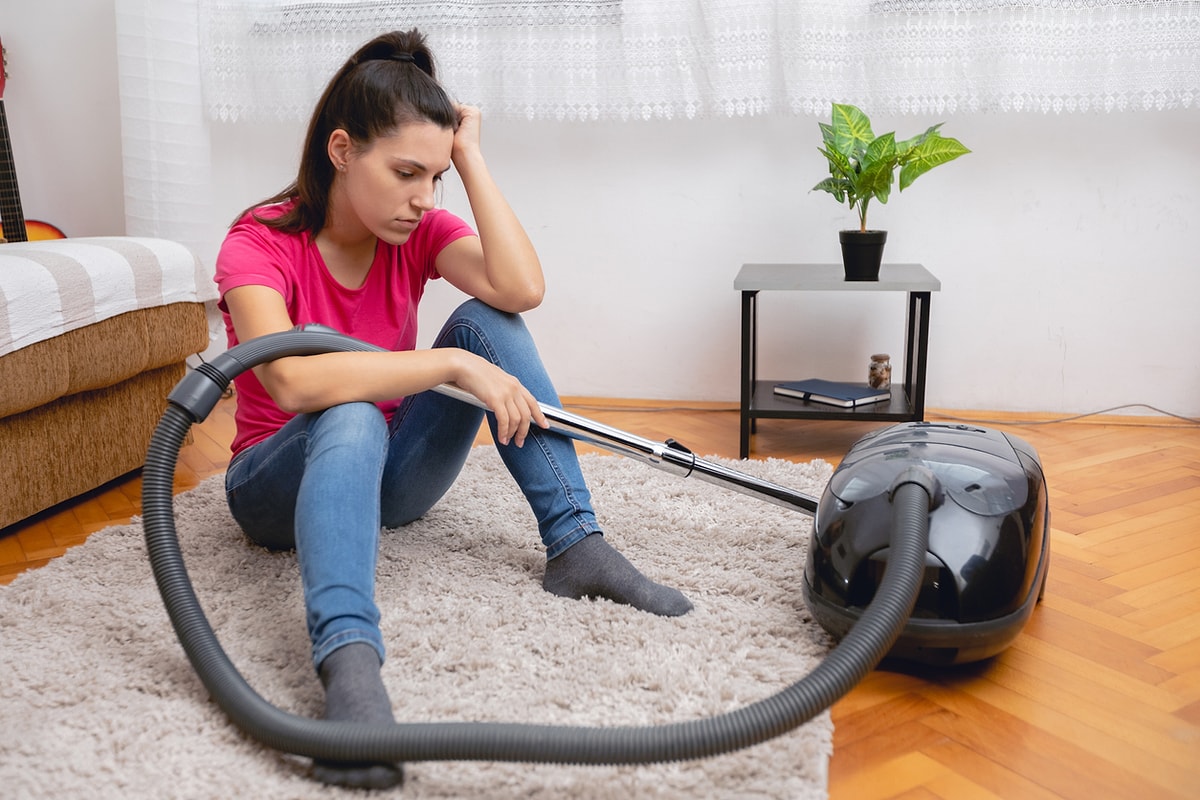 Flickan sitter på mattan och bredvid henne står en dammsugare.