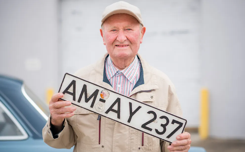 En äldre man håller upp sin bilnummerplåt, som han har haft sedan han köpte bilen.